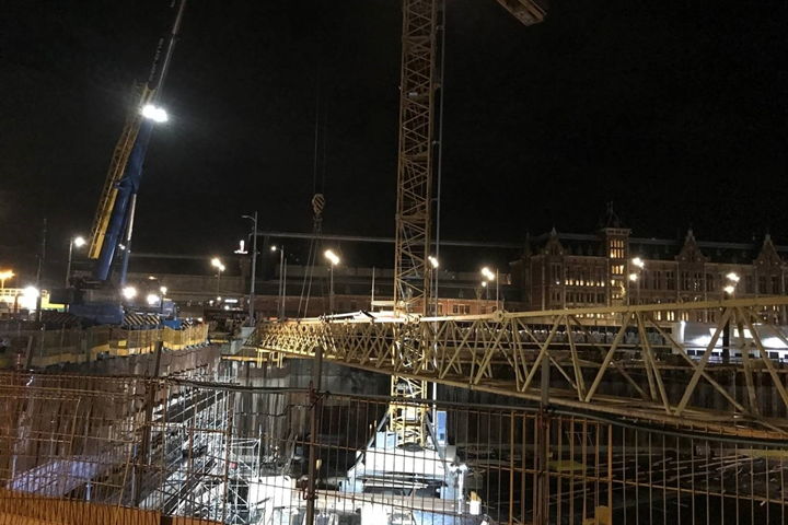 De torenkraan wordt ingehesen en opgebouwd aan de rand van de bouwkuip waardoor een kleinere mobiele kraan volstaat. Op de achtergrond Amsterdam Centraal.